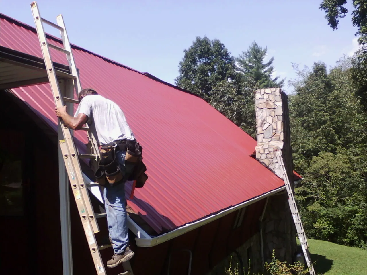 Roofer on a ladder beside a red metal roof and stone chimney — Soffit & Fascia Repair by Roofing Brothers in Chesapeake Beach